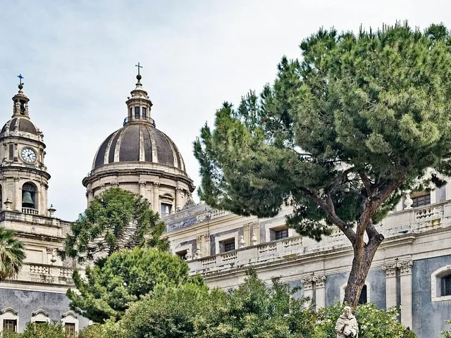 Ein Blick auf die Kuppel und den Glockenturm der Kathedrale von Catania hinter grünen Bäumen.