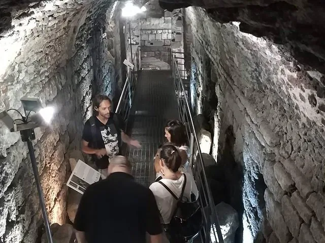 Eine Reisegruppe geht auf einem Metallsteg durch einen beleuchteten Steintunnel.